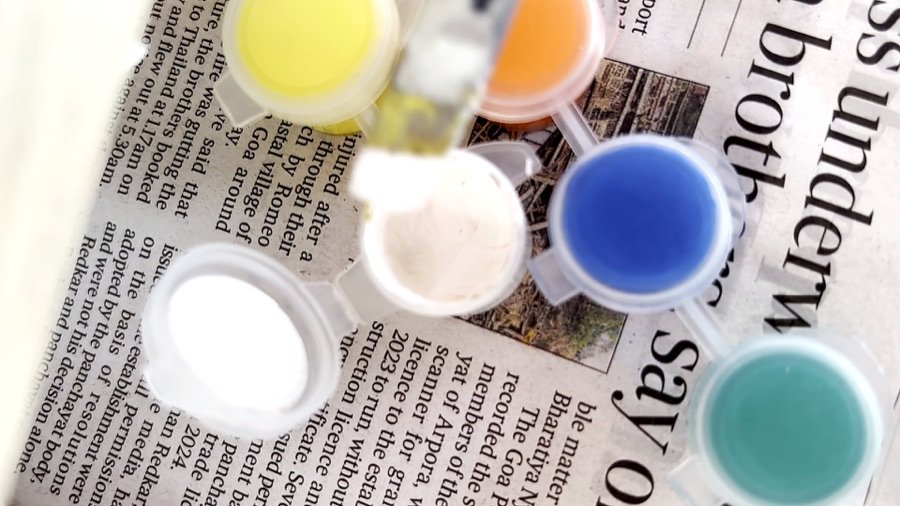 Small paint pots — blue, yellow, orange, white and teal — sitting on newspaper next to brushes.